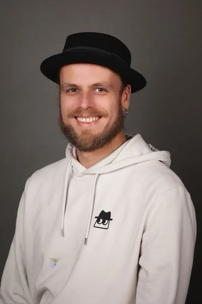 Mathieu Ritter smiling in a black hat and white hoodie, studio portrait against a grey background