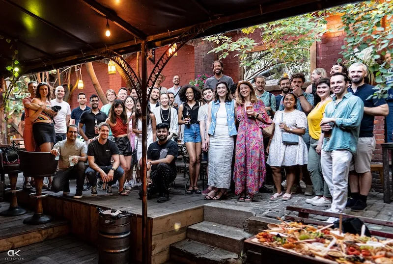 Large group of about 30 people posing together in a leafy backyard courtyard with string lights
