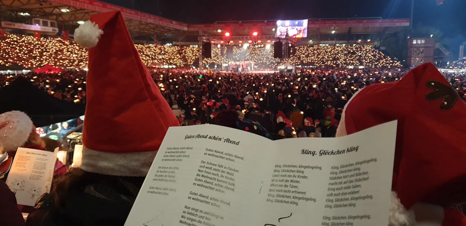 Thousands of fans holding candles at FC Union Berlin's Weihnachtssingen, with a Christmas carol song sheet in the foreground