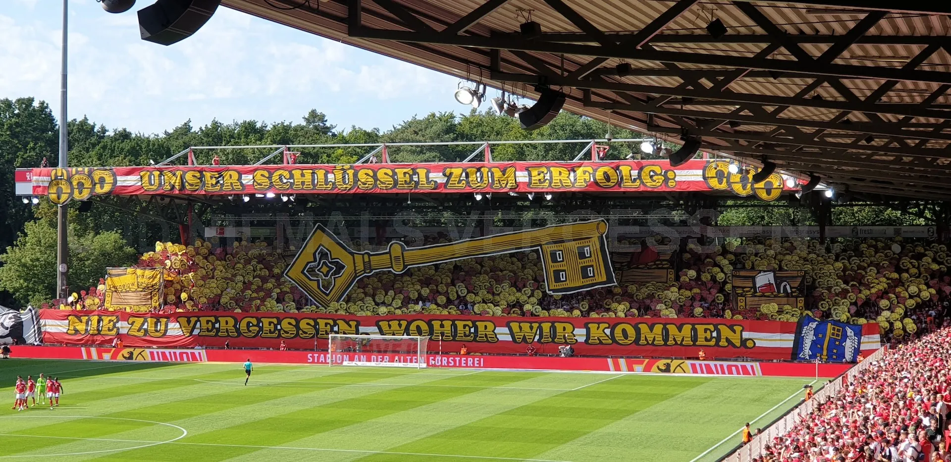 FC Union fans displaying a massive tifo at Alte Försterei reading "Unser Schlüssel zum Erfolg - Nie zu vergessen woher wir kommen" with a golden key