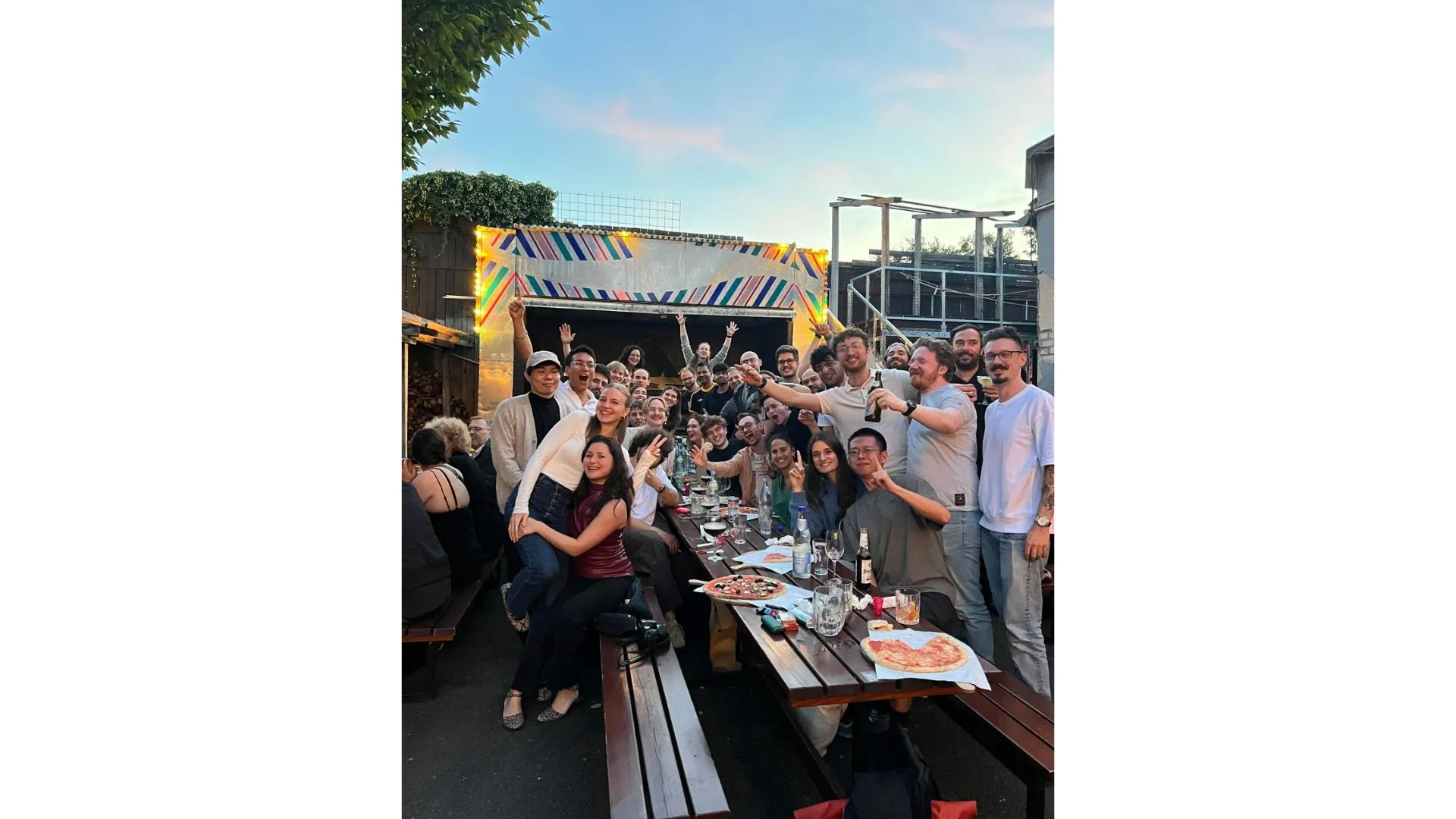 Almedia team gathering at an outdoor beer garden, about 20 people raising drinks at a long wooden table at sunset