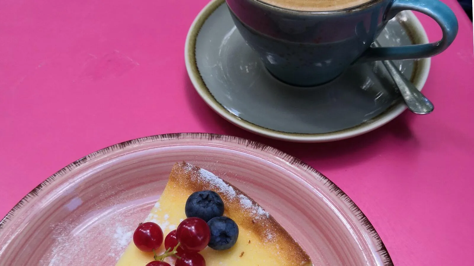 A cup of coffee and a slice of cheesecake topped with blueberries and red currants on a bright pink table